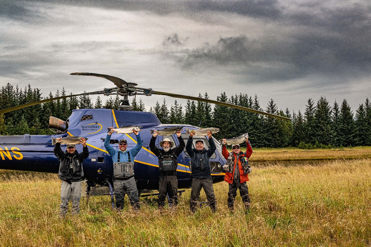 Five anglers holding salmon overhead in front of a Yakutat Expeditions helicopter in a grass field.